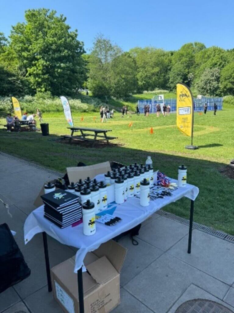 A table full of Bring the Power Merch in the foreground including waterbottles and notebooks with kids playing in the background on grass.