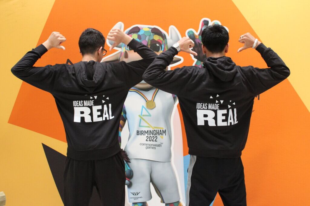 Two young people, backs facing the camera. Both people are pointing to the Ideas Made Real logo displayed on the back of their hoodies.